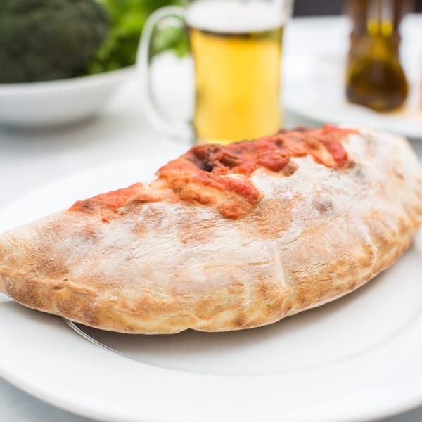 A calzone with tomato sauce on a white plate, with a beer and salad in the background.