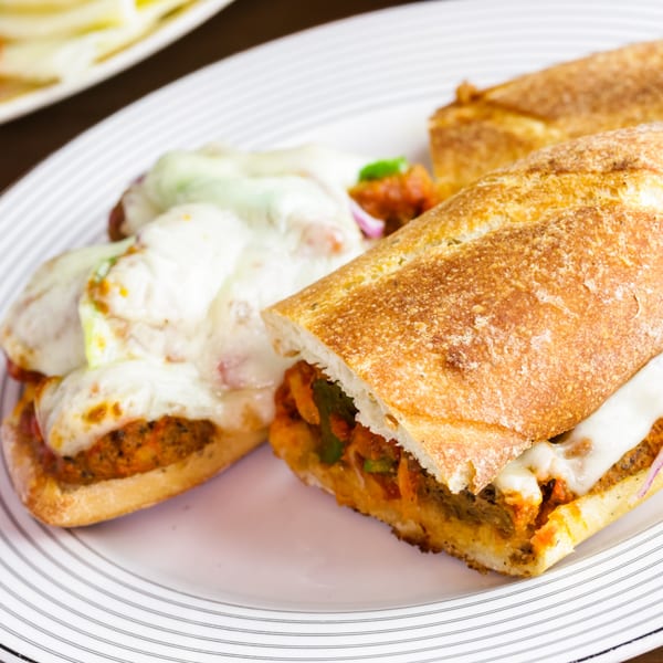Two cheesy meatball subs on a white plate, one sliced open revealing the filling.