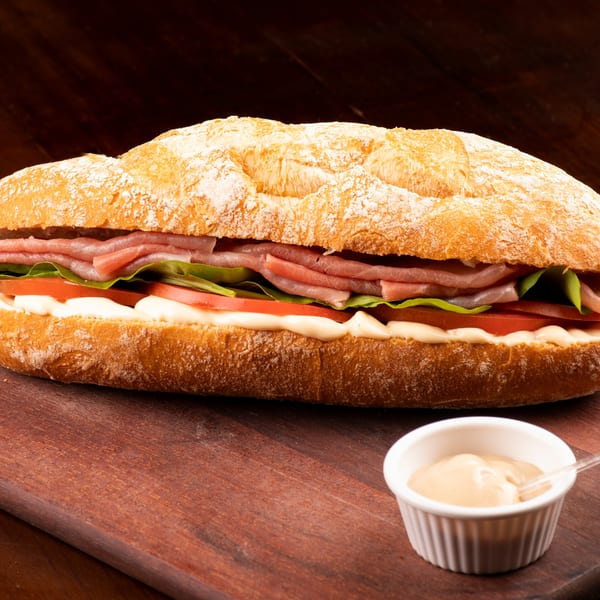 A long submarine sandwich with ham, lettuce, tomatoes, and mayonnaise, served with a side of sauce.